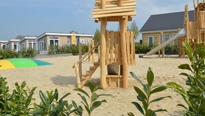Una estructura de juego de madera en una zona arenosa, con un edificio al fondo.