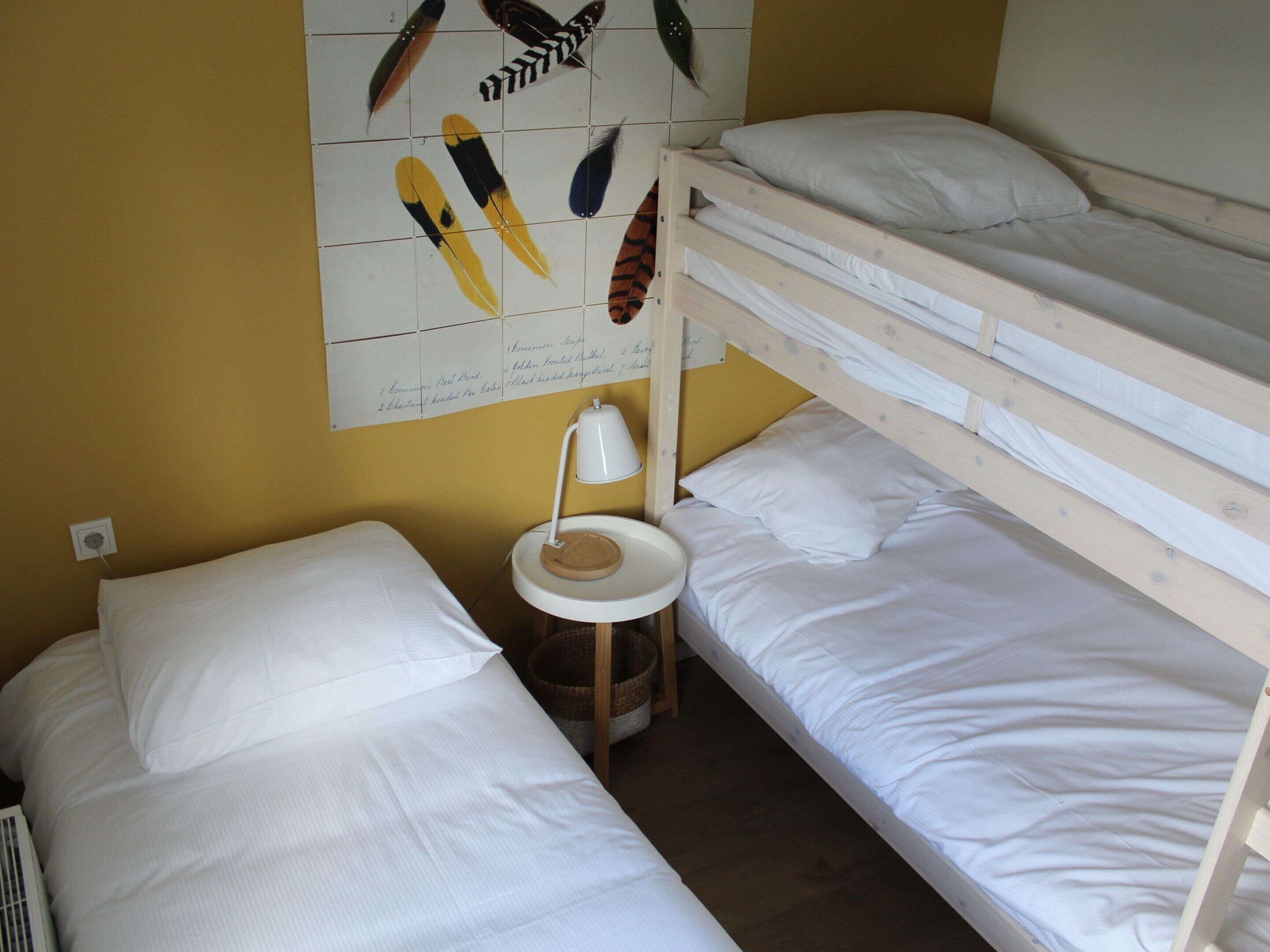 Una habitación pequeña y funcional con dos literas, una cama individual, una mesita con lámpara y un adorno de pájaro colgado en la pared.