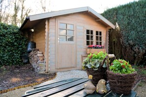 Una cabaña de madera con ventana, puerta y un pequeño jardín con plantas en macetas y una figurita de pájaro.