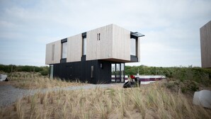 A modern building with a wooden exterior and a black lower section, situated on a sandy area with tall grasses. - Modern, Trendy F1 Theme Lodge, Near the Sea (Zandvoort)