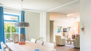 A modern living room with a dining area, featuring a wooden table, white chairs, and a large window with blue curtains. - Holiday Home on Texel With Spacious Garden (De Koog)