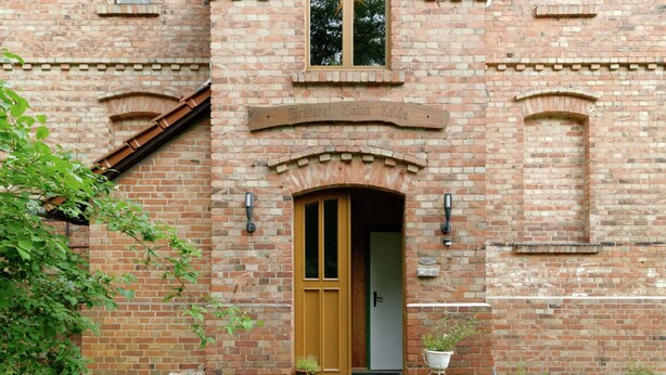 A brick building with a wooden door and a small porch area with a wheelbarrow and potted plants. - Flat in Furstenwald Near Berlin Forest (Fürstenwalde/Spree)