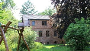 A brick building with a tiled roof, surrounded by lush greenery and a swing set in the foreground. - Flat in Furstenwald Near Berlin Forest (Fürstenwalde/Spree)