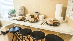 Un comptoir de cuisine avec une machine à café, des tasses et des pâtisseries. Il y a quatre tabourets et une poubelle.