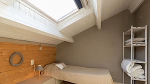 A small, cozy bedroom with a sloped ceiling, a wooden headboard, a single bed, a bedside table with a lamp, a shelf with folded towels, and a skylight. - Beautiful Cottage in Malempre With Fenced Garden (Manhay)