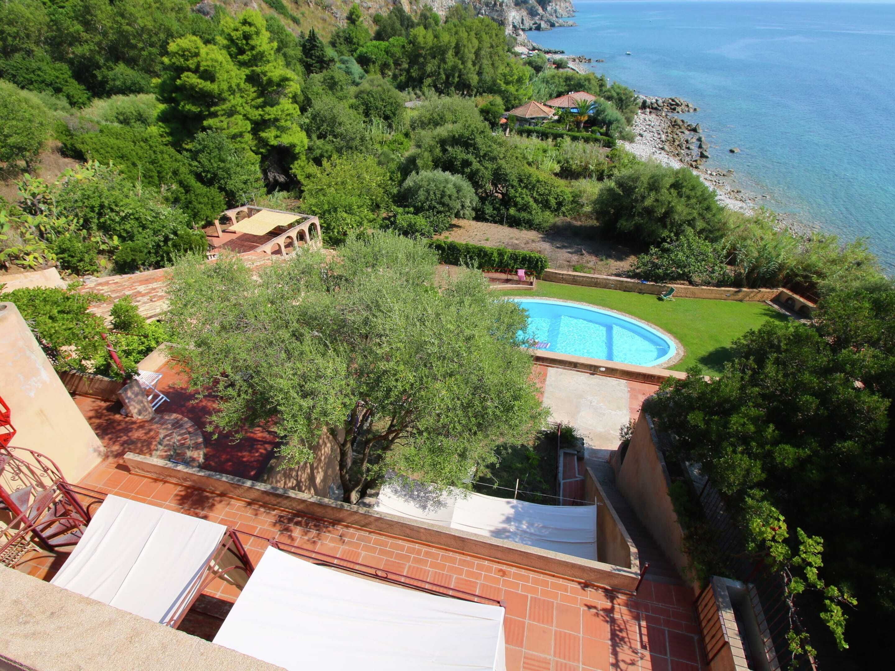 A rooftop pool with a view of the sea, surrounded by lush greenery and a small building with a red roof.