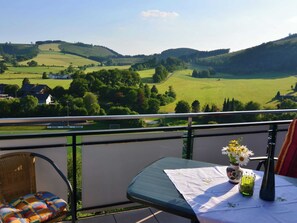 A balcony with a wicker chair, a small table with a vase of flowers, and a view of green fields and hills. - Ruhige Wohnung in Meschede im Sauerland mit Balkon (Meschede)