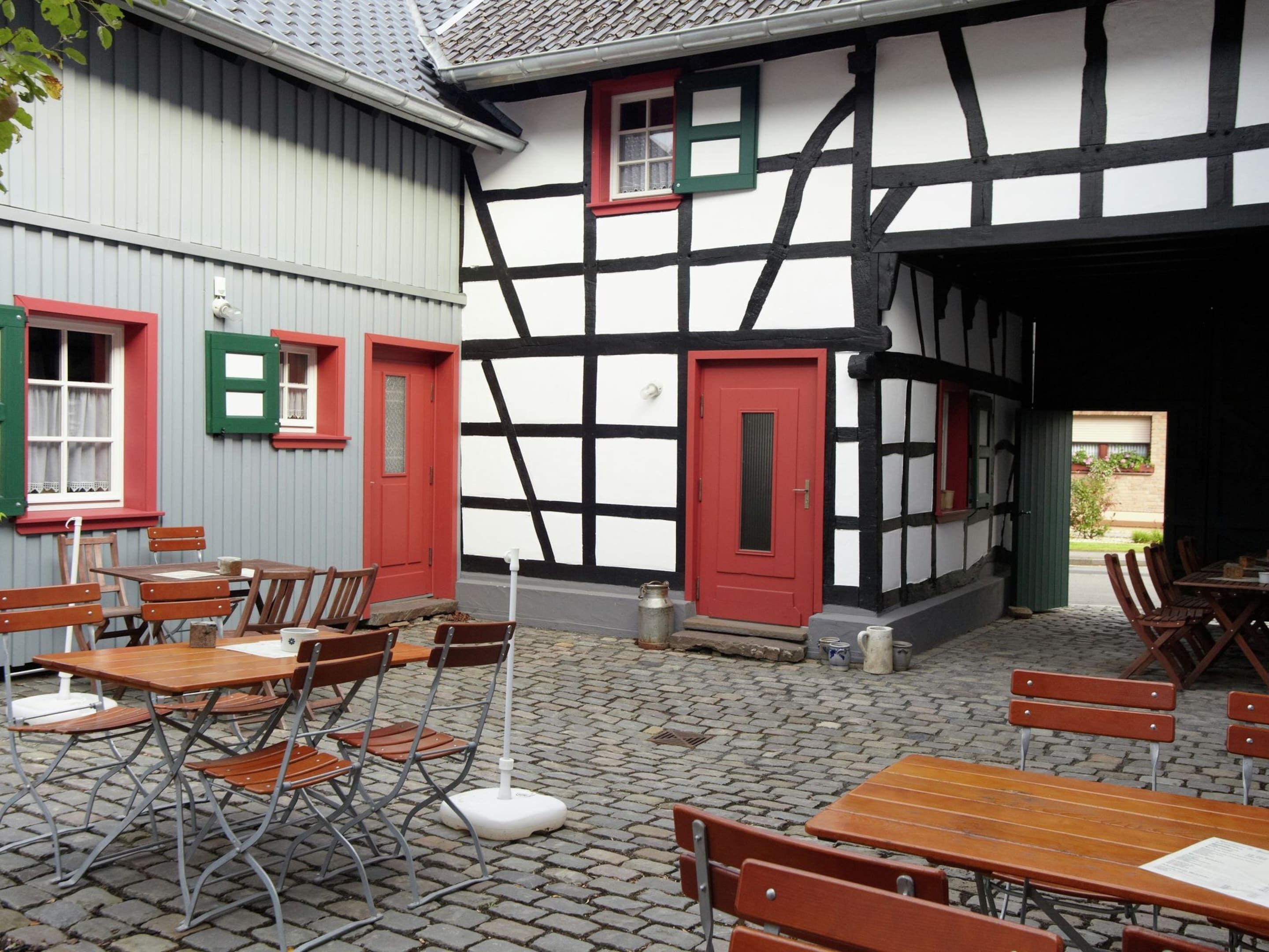 A traditional half-timbered building with a red door and windows, outdoor seating area with wooden tables and chairs, and a cobblestone courtyard.