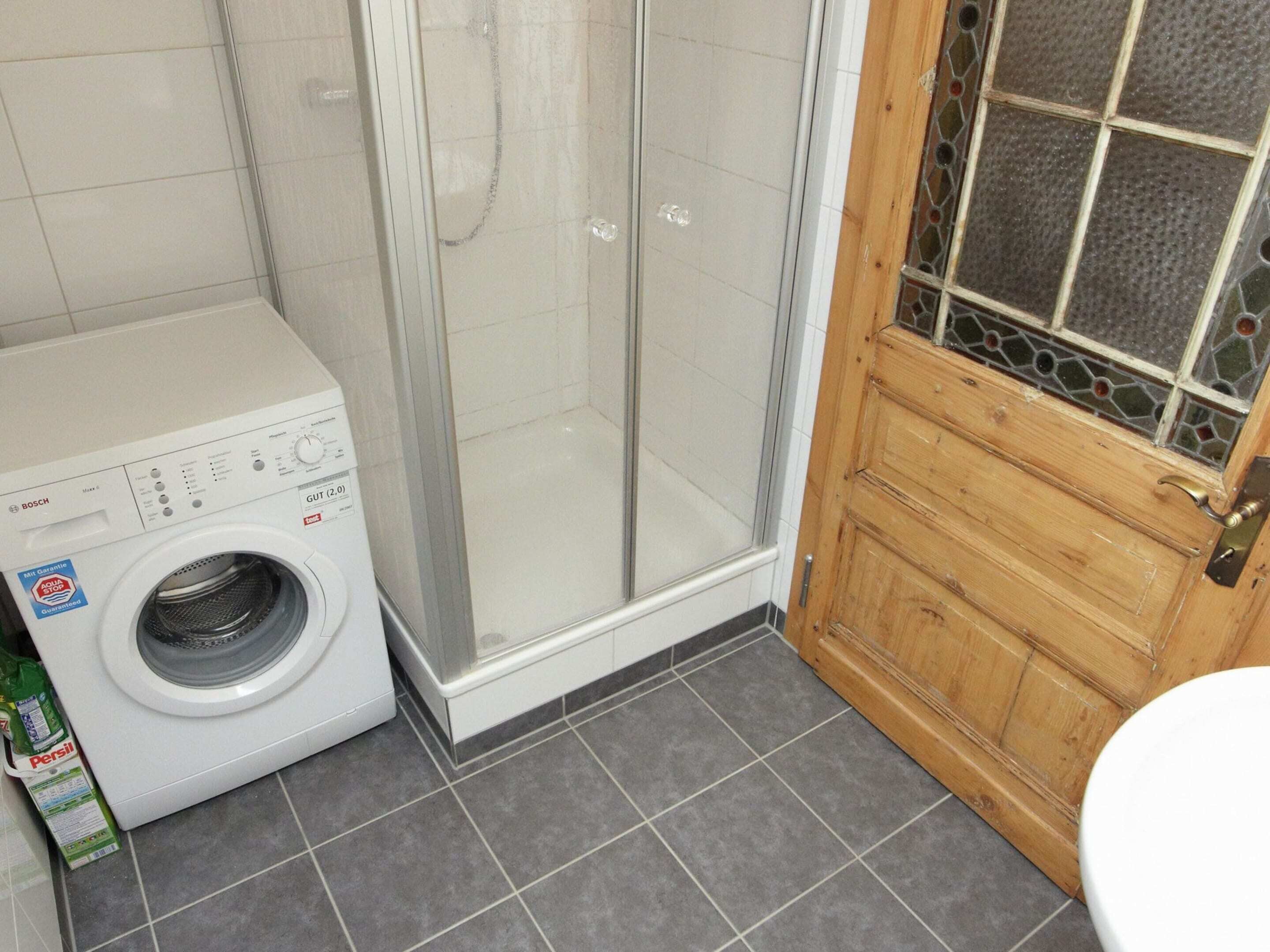 A compact bathroom with a washing machine, a shower enclosure, and a wooden door with a glass window.