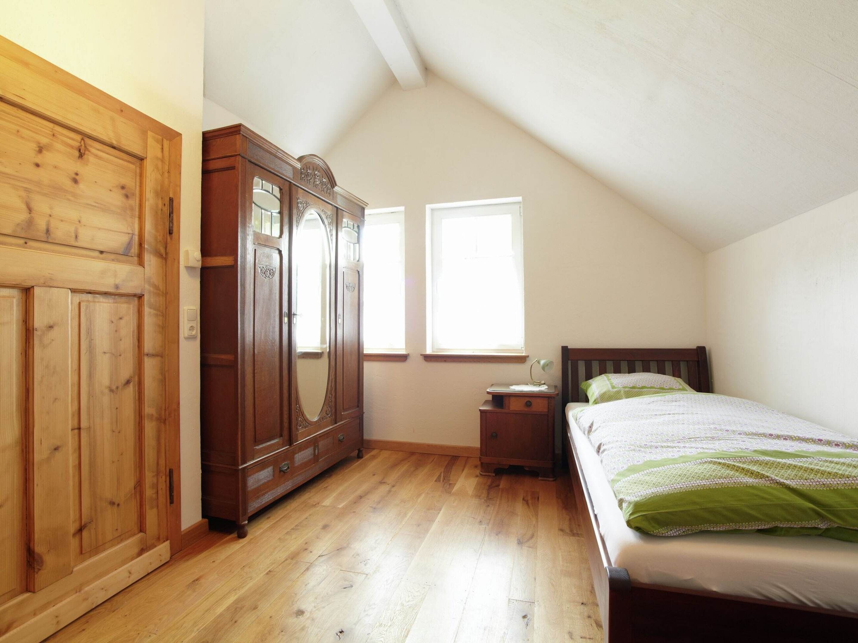 Un dormitorio con un armario de madera, una cama con ropa de cama verde, una mesita de noche y una ventana que proporciona luz natural.