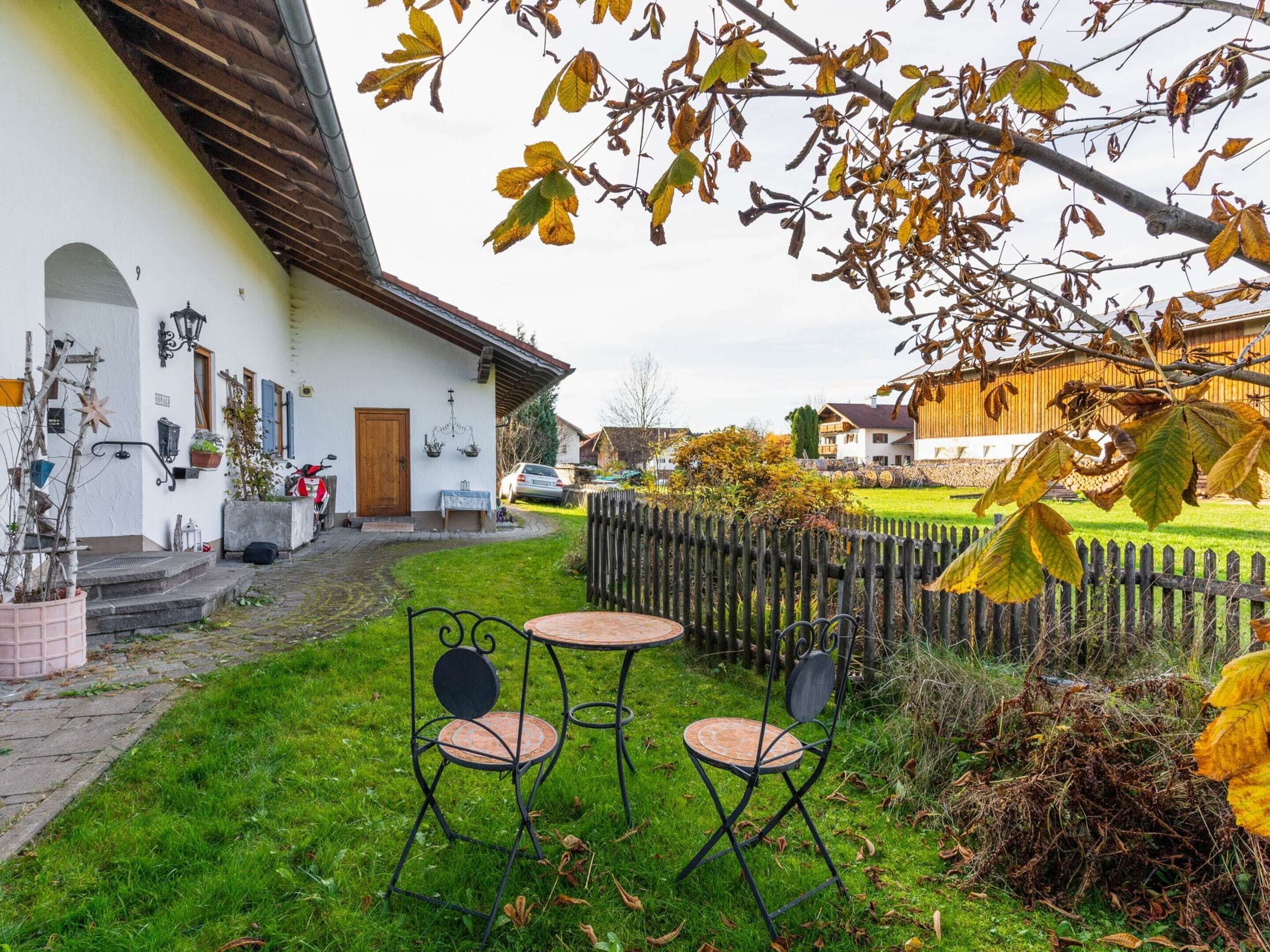 Une cour avec un bâtiment blanc, une clôture en bois et un petit jardin agrémenté de deux chaises en métal et d’une table.