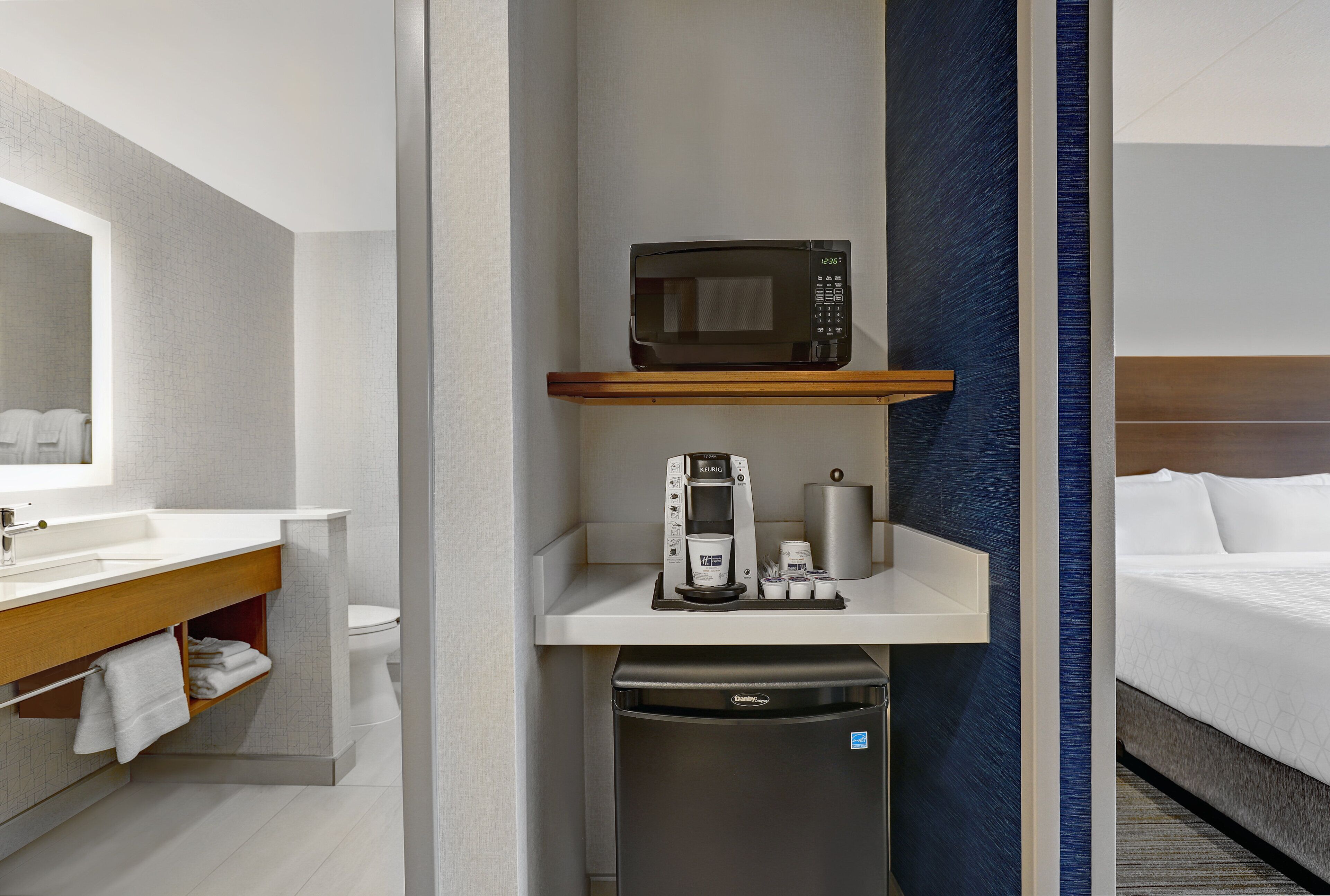 Une chambre d’hôtel compacte avec un lavabo blanc, un four à micro-ondes posé sur une étagère, une machine à café, un petit réfrigérateur et un lit avec du linge de lit blanc.