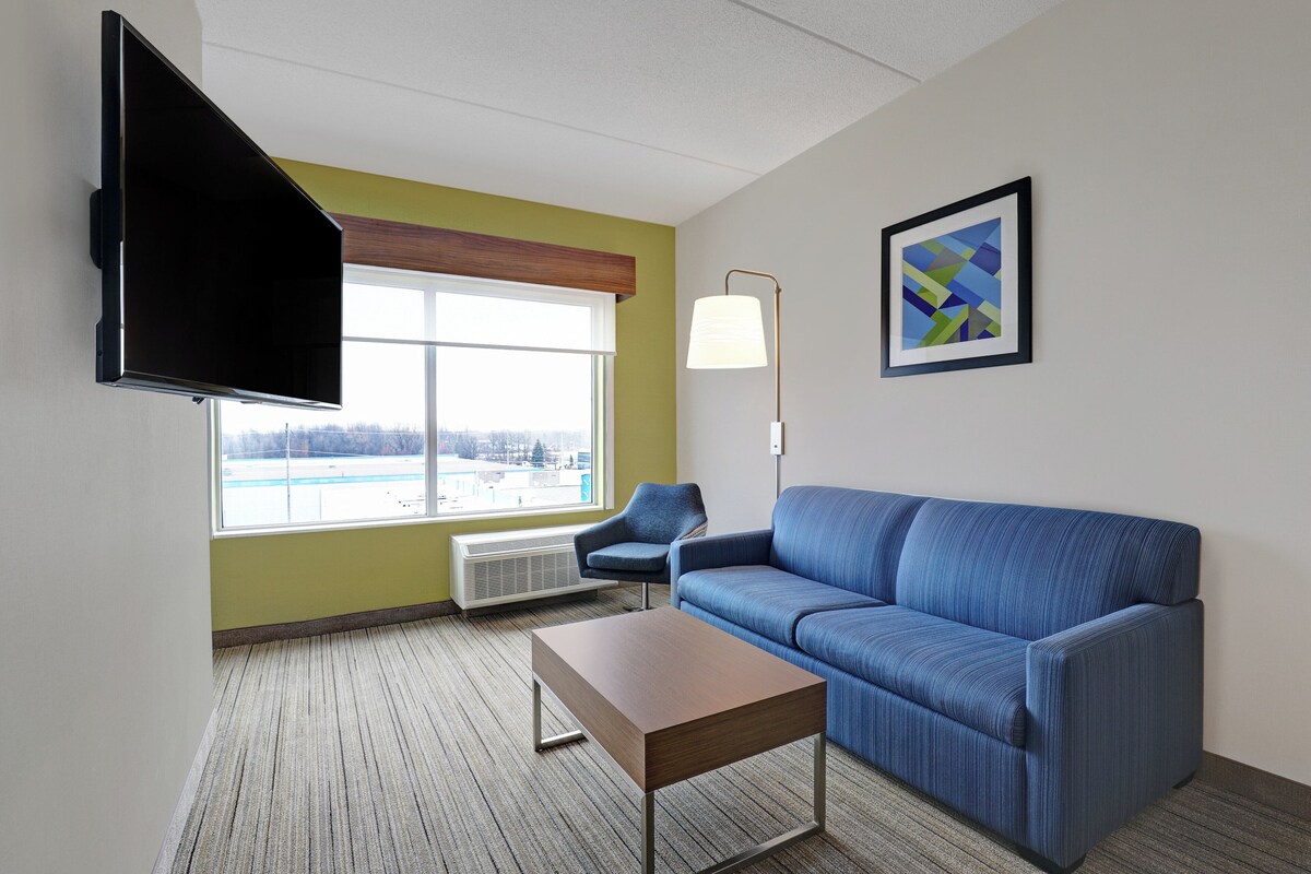 A modern hotel room with a blue sofa, a wooden coffee table, a flat-screen TV mounted on the wall, a floor lamp, and a large window offering a view of the outdoors.