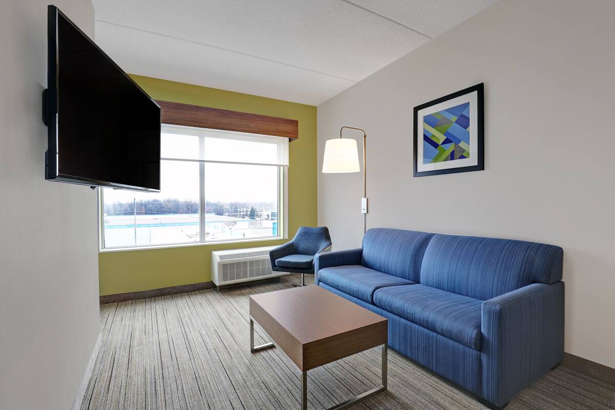 Une chambre d’hôtel moderne avec un canapé bleu, une table basse en bois, un téléviseur à écran plat fixé au mur, une lampe sur pied et une grande fenêtre donnant sur l’extérieur.