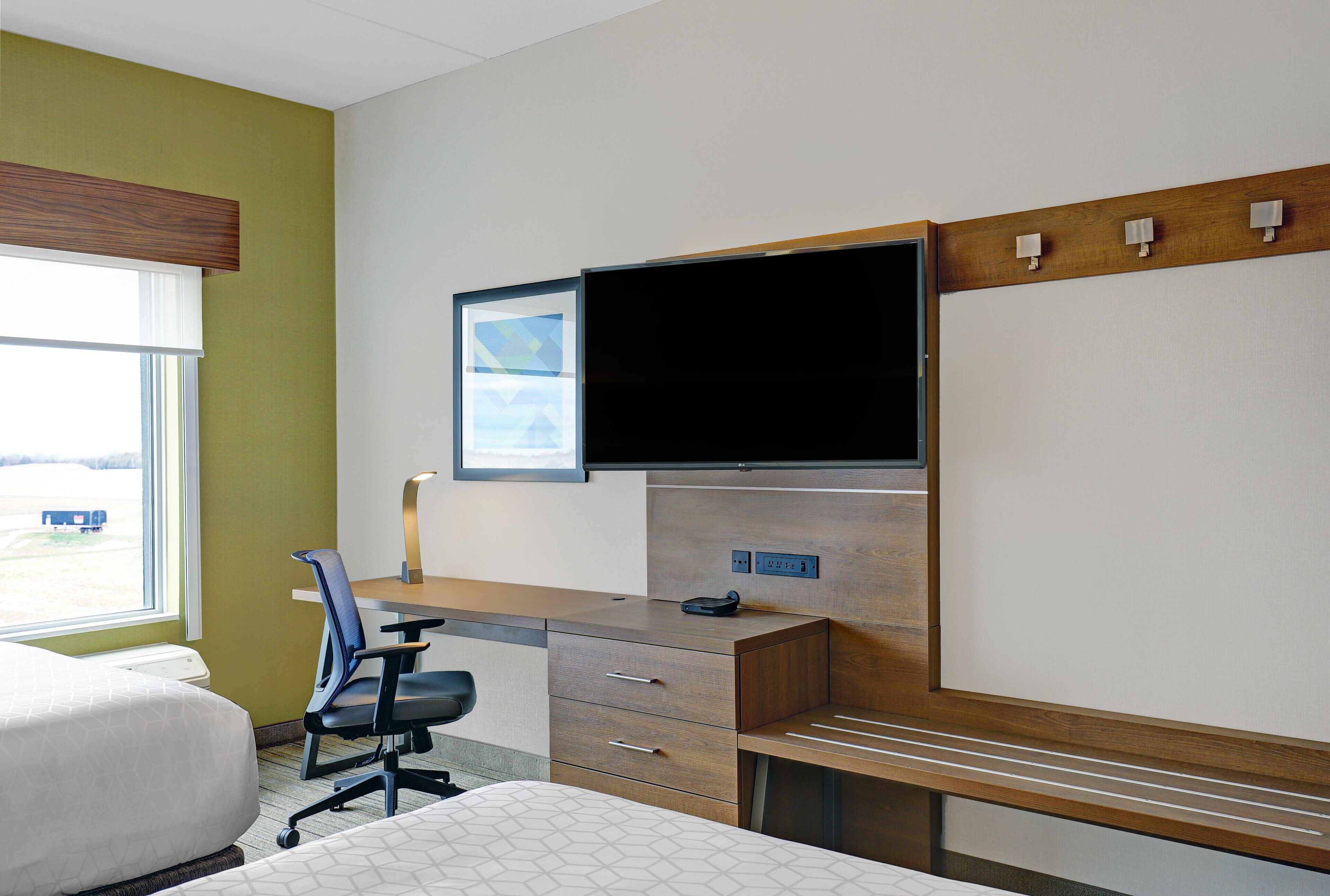 Une chambre d’hôtel comprenant un lit, un bureau avec une chaise, un téléviseur à écran plat fixé au mur et une fenêtre avec des stores.