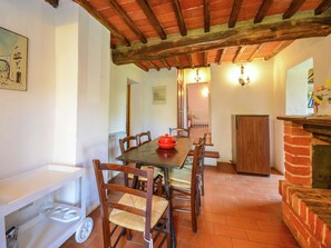 A dining area with a wooden table, chairs, and a red bowl. There is a brick fireplace and a white shelf with a kettle. - Cozy Farmhouse With Pool (Castiglione del Lago)