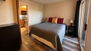 Ein Schlafzimmer mit Bett, Nachttisch, Kommode und Blick auf eine Küche durch eine offene Tür.