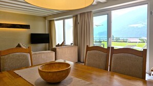 A dining area with a wooden table, chairs, a wooden bowl, a flat-screen TV, and a large window with a view of the outdoors.