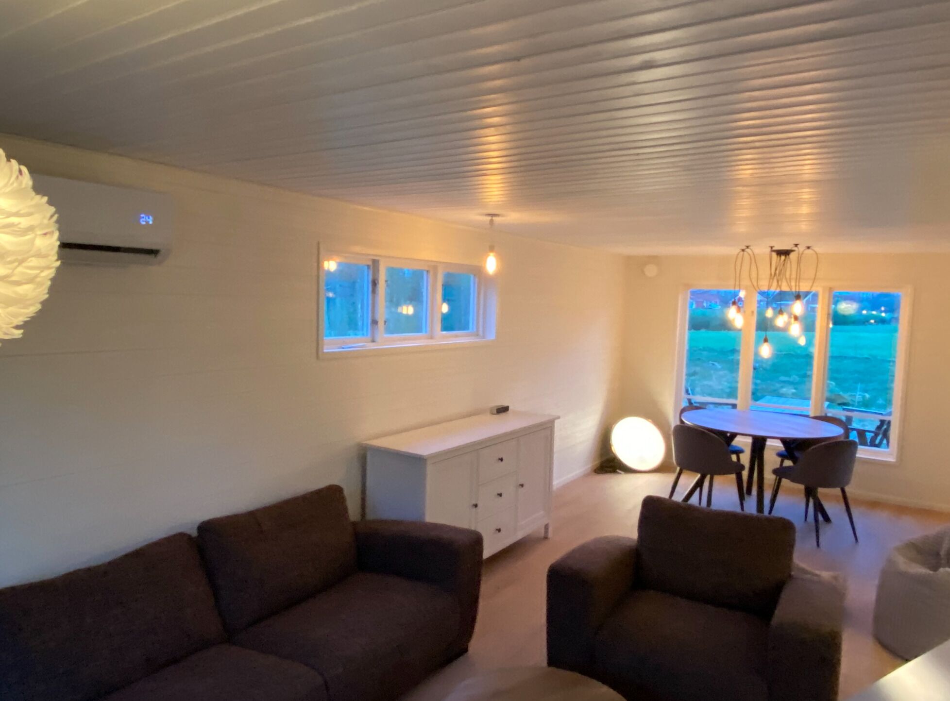 Ett modernt vardagsrum med en soffa, ett matbord med stolar, ett vitt skåp och ett stort fönster med utsikt över havet.