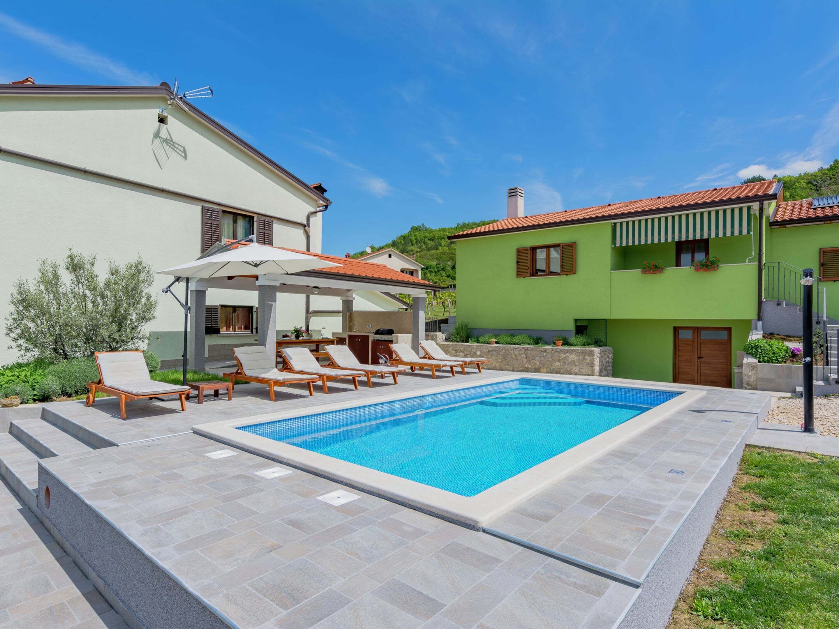 Une piscine entourée de chaises longues et d’un espace de repos couvert, le tout bordé d’une maison à deux étages aux murs verts et au toit recouvert de tuiles rouges.