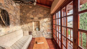 A stone-walled room with a wooden ceiling, a wooden bench, a wooden table, and a large window with a view of a hillside. - Traditional Apartment in Elounda (Agios Nikolaos)