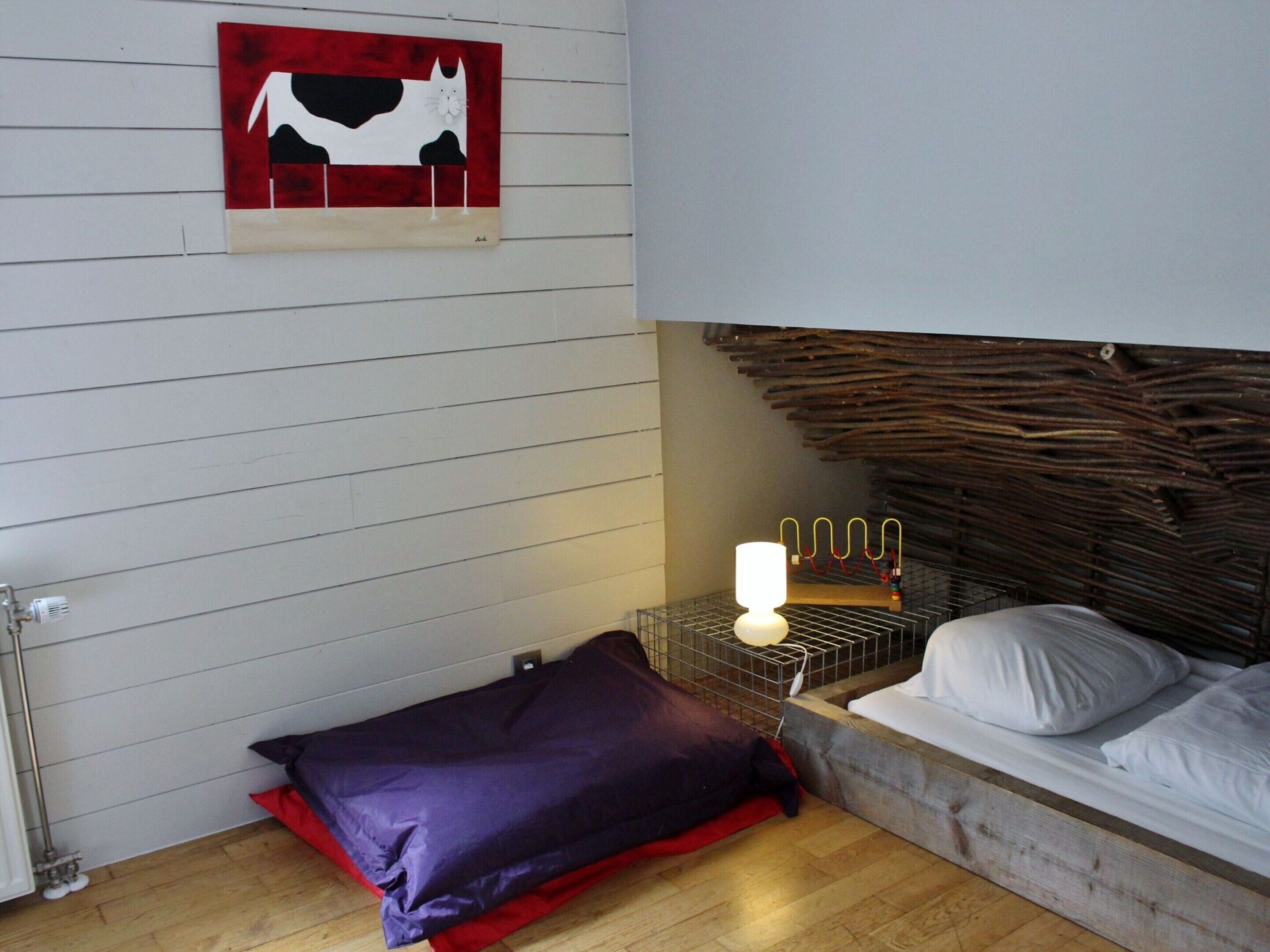 A bedroom with a bed, a headboard, a wall-mounted artwork, a bedside lamp, and a red and purple pillow.