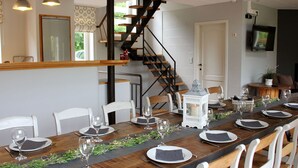 A dining area with a long wooden table set for a meal, white chairs, and a staircase in the background. - Holiday Home in Jupille Near Forest Trails (Rendeux)