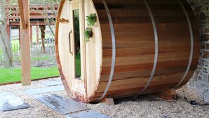 A wooden barrel-shaped sauna with a door and a small window, set on a gravel surface with a wooden deck in the background. - Holiday Home in Jupille Near Forest Trails (Rendeux)