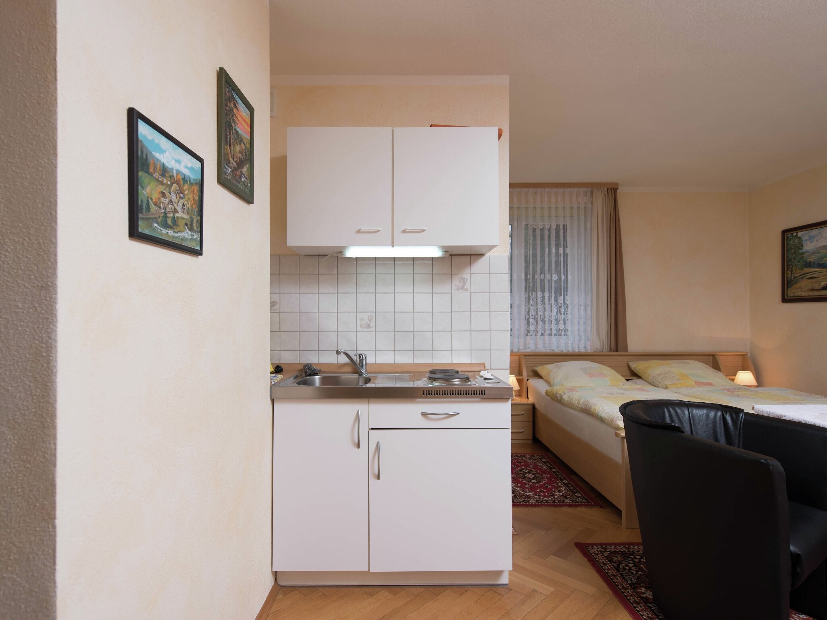 A compact room with a kitchenette, a bed with a headboard, a nightstand, and a chair.
