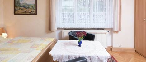 Un dormitorio con cama, una mesa con un florero, una silla, un radiador y una ventana con cortinas.