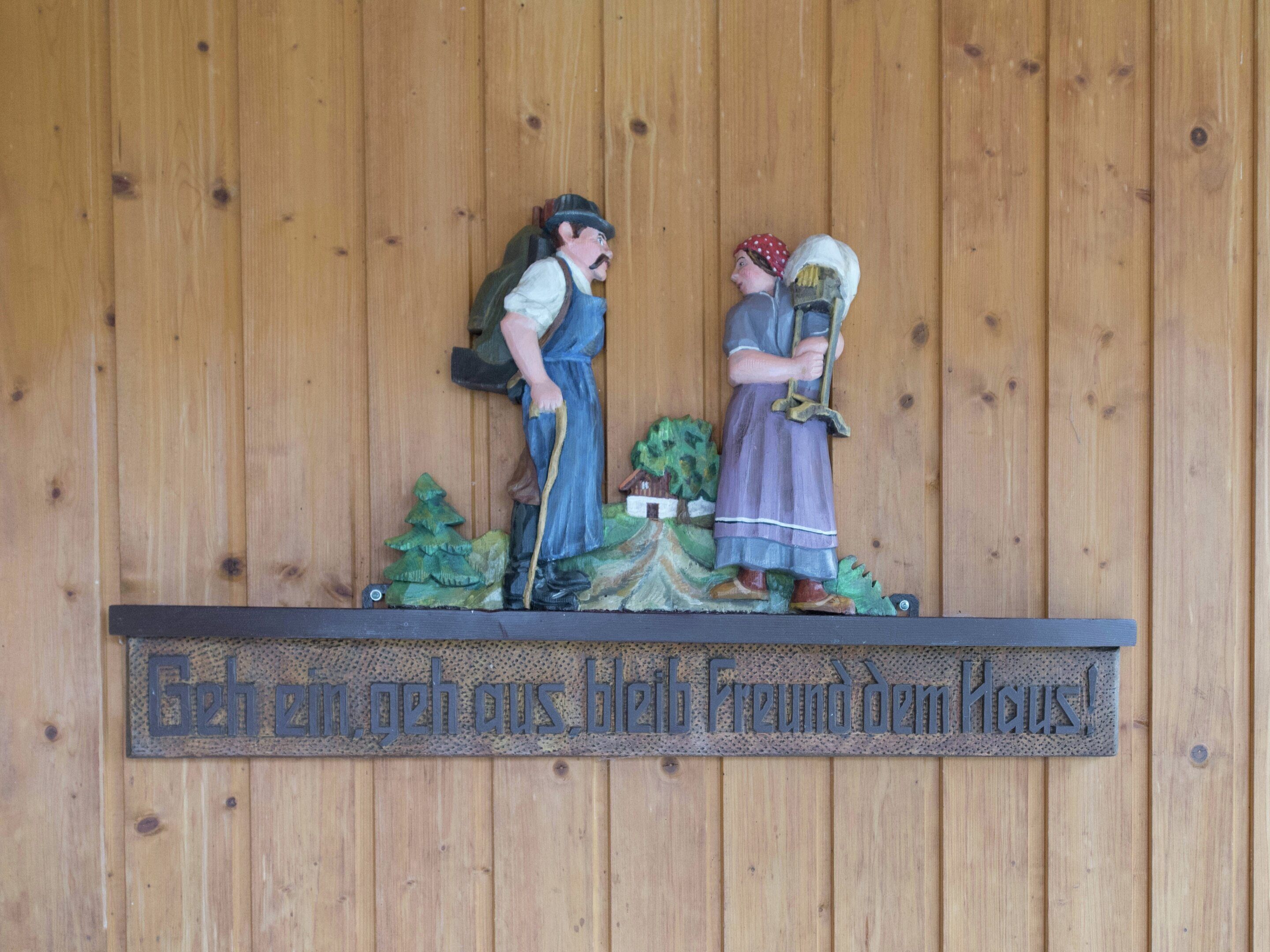 A wooden wall with a plaque that reads "Geh ein, geh aus. Bleib Freund dem Haus!" and a figurine of a man and woman.