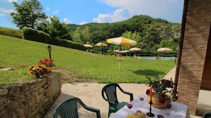 Un patio al aire libre con una mesa dispuesta para una comida, rodeado de sillas verdes y con vistas a una piscina y colinas al fondo.
