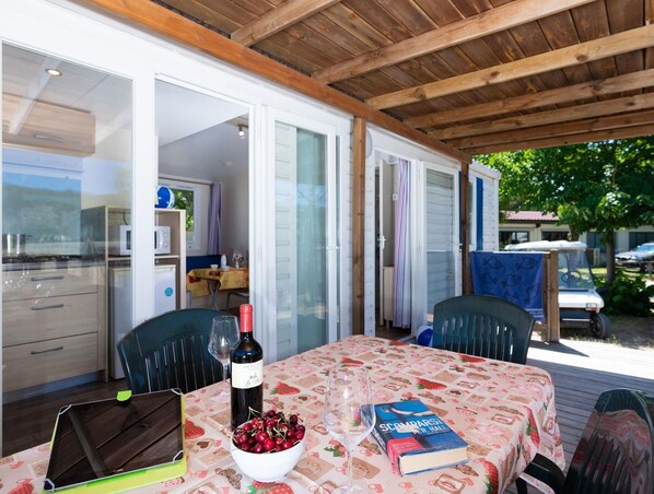 An outdoor dining area with a table set for two, a laptop, a bowl of cherries, a wine bottle, and a book.
