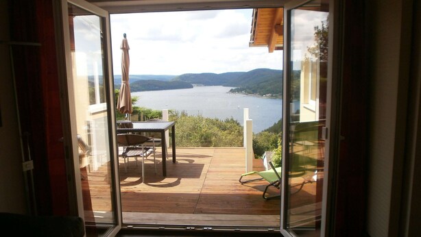 A room with a sliding glass door leading to a deck with a table and chairs, overlooking a lake and hills. - Luxurious Apartment in Waldeck With Sauna (Waldeck)