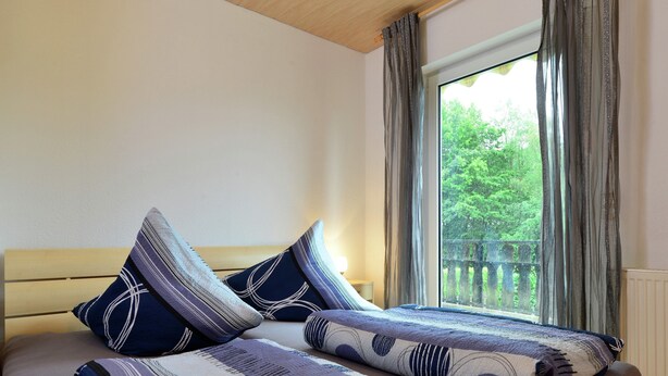 Ein Schlafzimmer mit einem Holz-Kopfteil, einem großen Fenster mit Vorhängen und Blick ins Grüne.
