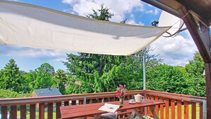 A covered outdoor area with a picnic table, a small table with a vase and a teapot, and a view of a green landscape.