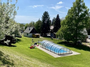 Ein Schwimmbecken mit Glasdach, umgeben von einer Rasenfläche und Bäumen.