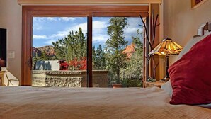 A bedroom with a large bed, a view of trees and mountains through a sliding door, and a lamp on the side table. - Stunning Luxury Home, Amazing Views! Painted Cliff (Sedona)