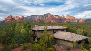 A two-story house with a tiled roof, surrounded by trees and red rock formations in the background. - Stunning Luxury Home, Amazing Views! Painted Cliff (Sedona)