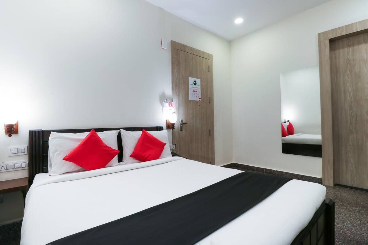 Hotellihuone, jossa on sänky, puuovi, peili ja pieni pöytä, jonka päällä on punainen tyyny.