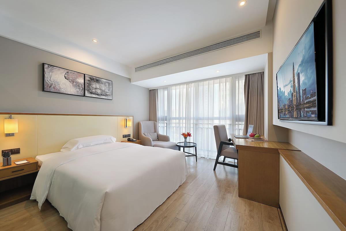 Ein Hotelzimmer mit einem großen Bett, einem Schreibtisch mit einem Laptop, zwei Stühlen, einem Gemälde und einem großen Fenster mit Vorhängen.