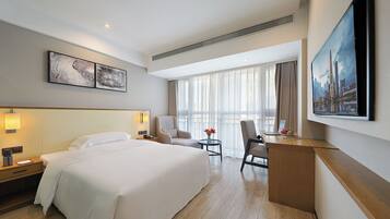 Quarto de hotel com uma cama grande, uma escrivaninha com um laptop, duas cadeiras, um quadro e uma janela ampla com cortinas.