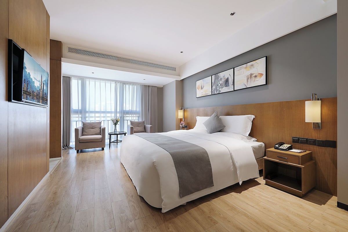 Ein modernes Hotelzimmer mit einem großen Bett, einem Schreibtisch, einem Stuhl und Blick auf die Stadt durch große Fenster.