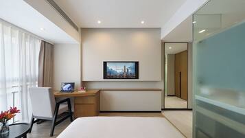 Una habitación de hotel moderna con cama, escritorio con computadora portátil, silla, mesita con flores, un cuadro de paisaje urbano en la pared y una división de vidrio.