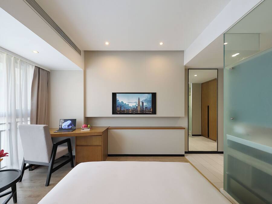 Una habitación de hotel moderna con cama, escritorio con computadora portátil, silla, mesita con flores, un cuadro de paisaje urbano en la pared y una división de vidrio.