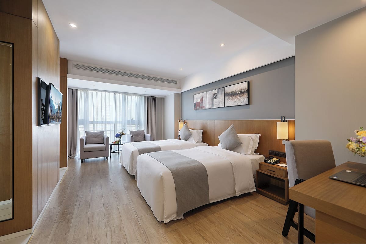 Ein Hotelzimmer mit zwei Betten, einem Schreibtisch, einem Sessel und einem Fernseher, mit Blick auf die Stadt.