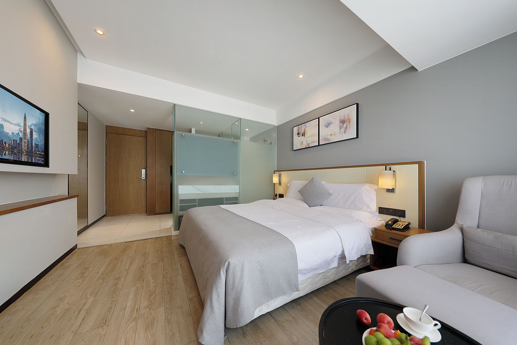 A hotel room with a bed, a sofa, a glass partition, a painting, and a table with fruits.