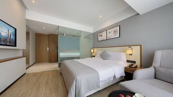 A hotel room with a bed, a sofa, a glass partition, a painting, and a table with fruits.
