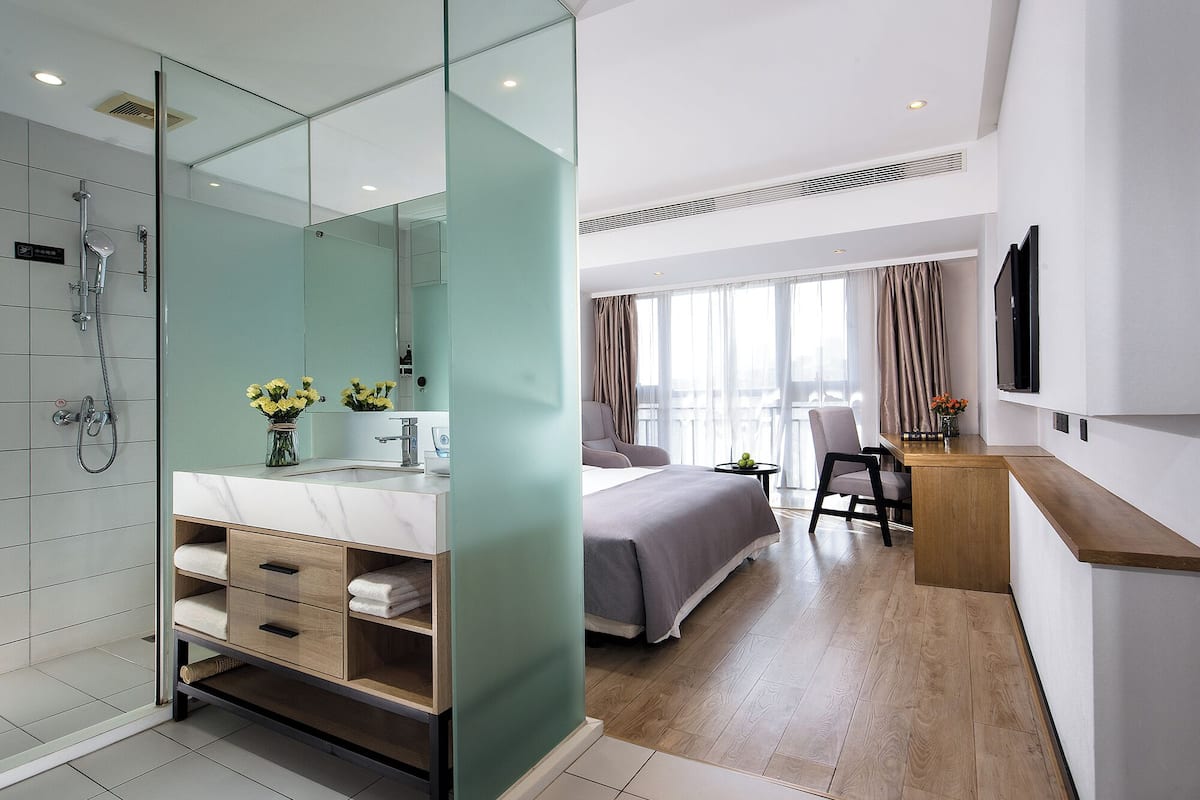 Ein modernes Hotelzimmer mit einer gläsernen Dusche, einem großen Bett, einem Schreibtisch mit Stuhl und einem an der Wand montierten Fernseher.