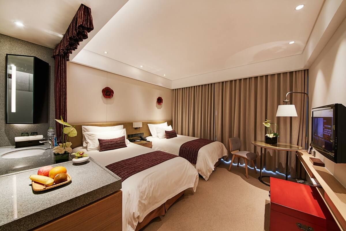 A hotel room with two beds, a desk with a lamp, a TV, a mirror, and a fruit bowl on the counter.